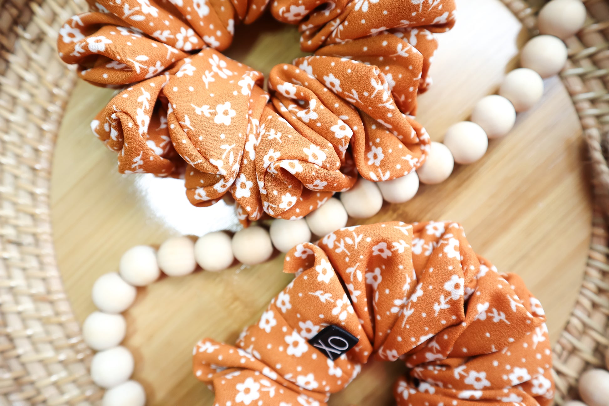 Brown scrunchies with white floral pattern on a wooden surface with white beads. Fabric might look brighter due to lighting.